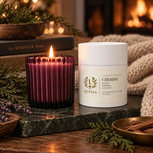 Lit Leveiza Cherish candle and white tube with evergreen sprigs, lavender, cinnamon sticks, a knitted blanket, and a glowing fireplace