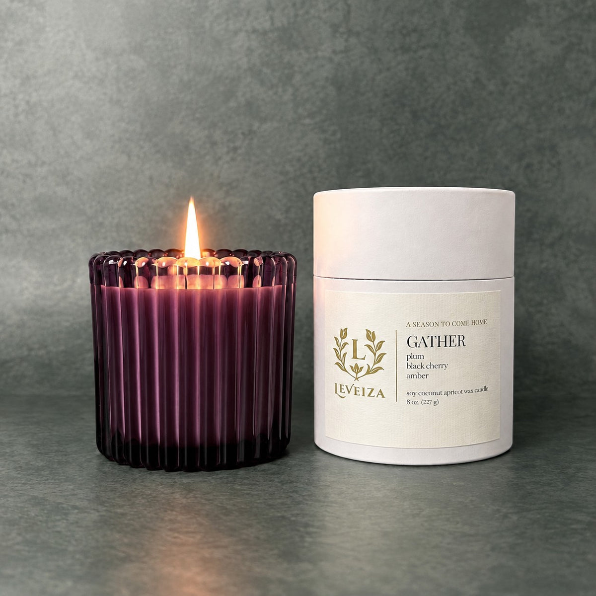 Lit Gather scented candle by Leveiza in deep plum ribbed glass beside its white labeled tube packaging