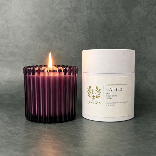Lit Gather scented candle by Leveiza in deep plum ribbed glass beside its white labeled tube packaging