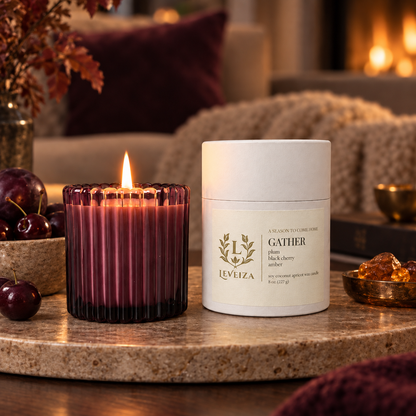 Lit Leveiza Gather candle and white tube in a cozy autumn setting with plums, cherries, knitted blankets, and a glowing fireplace