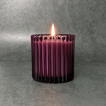 Lit candle in a deep plum ribbed glass vessel against a textured gray background.