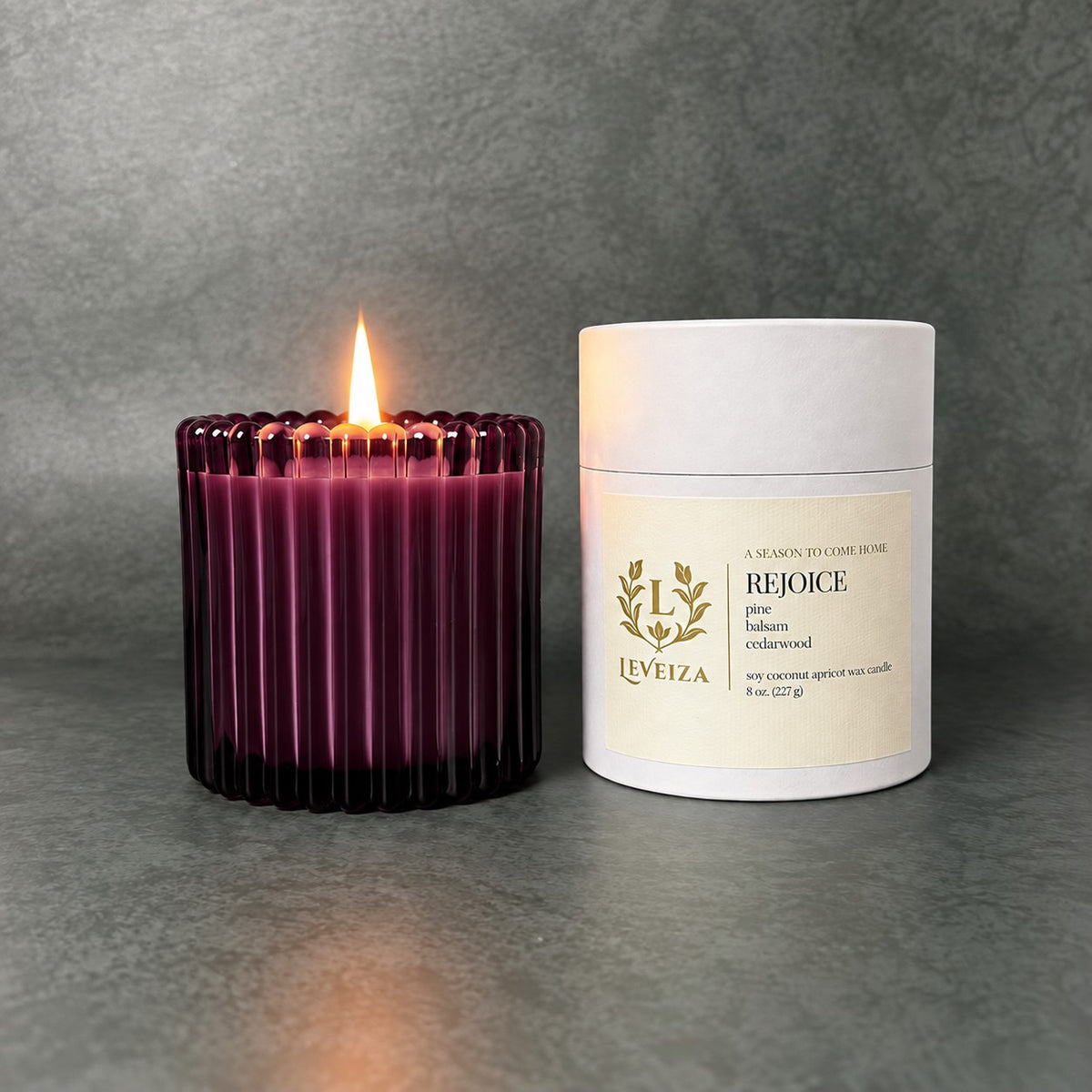 Lit Rejoice scented candle by Leveiza in deep plum ribbed glass beside its white labeled tube packaging