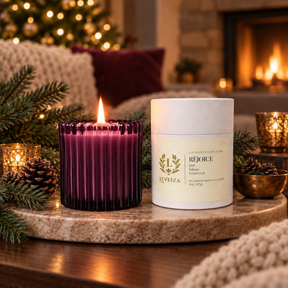 Lit Leveiza Rejoice candle and white tube with evergreen branches, pinecones, candlelight, a Christmas tree, and a glowing fireplace