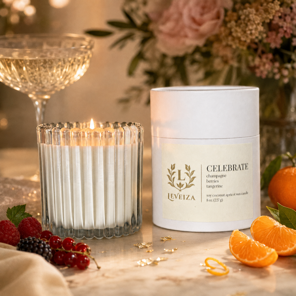 Lit Celebrate candle and tube surrounded by champagne, berries and tangerine in an elegant celebration setting.
