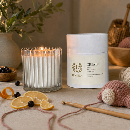 Lit Create candle and tube beside crochet work, citrus slices and black currants on a warm crafting table.
