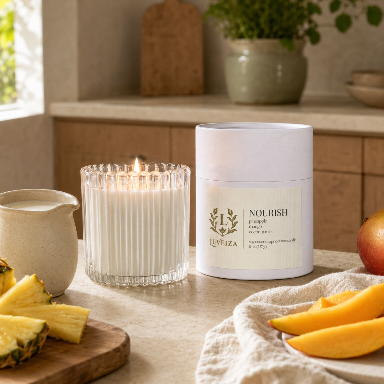 Lit Nourish candle and tube with pineapple, mango and coconut milk in a bright, welcoming kitchen.
