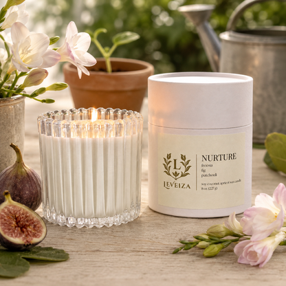 Lit Nurture candle and tube with freesia, ripe figs and a young seedling on a sunlit garden table.
