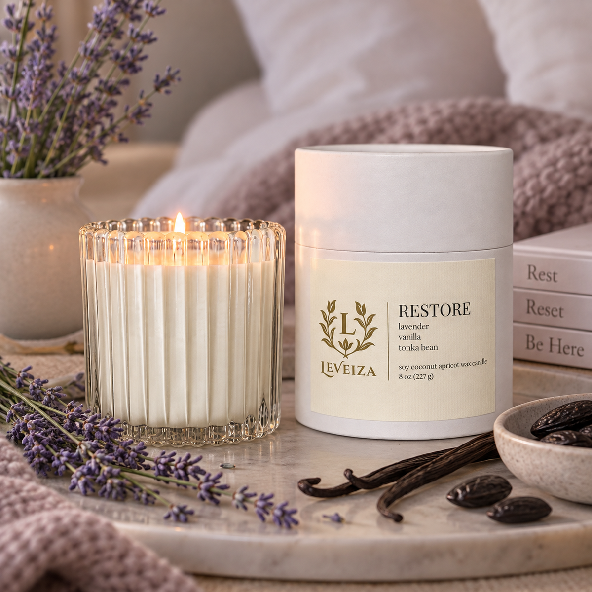 Lit Restore candle and white tube on a marble tray with lavender, vanilla pods, and tonka beans beside soft bedding