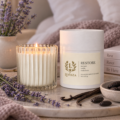 Lit Restore candle and white tube on a marble tray with lavender, vanilla pods, and tonka beans beside soft bedding