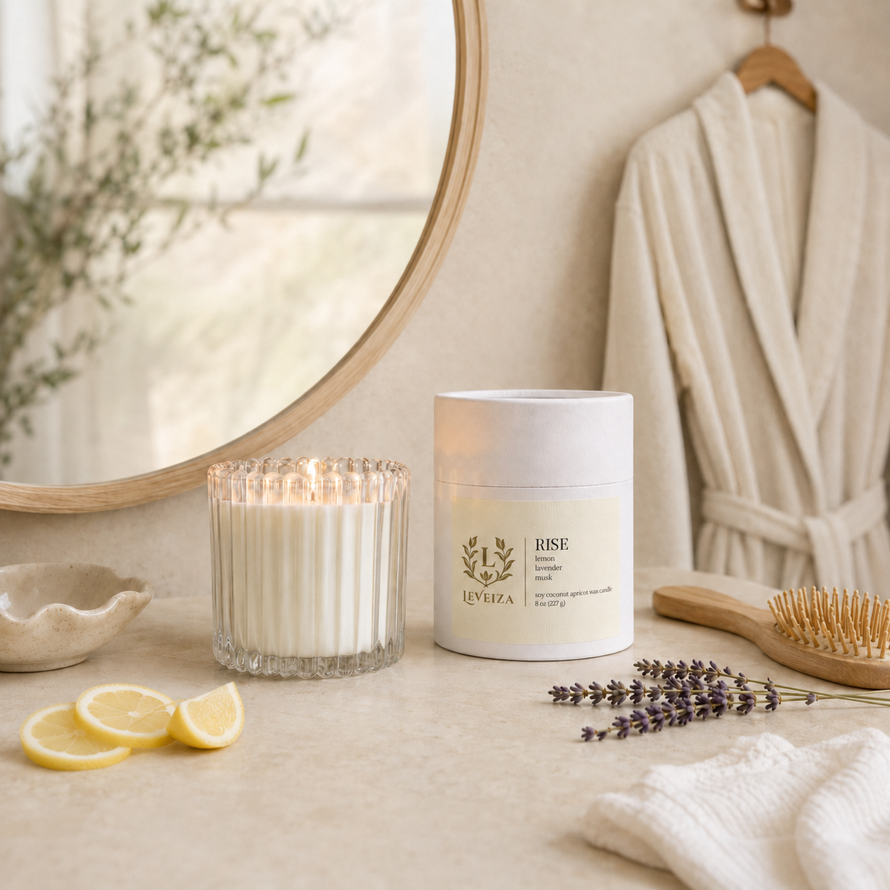 Lit Rise candle and tube with fresh lemons and lavender on a bright morning vanity with a robe and mirror.

