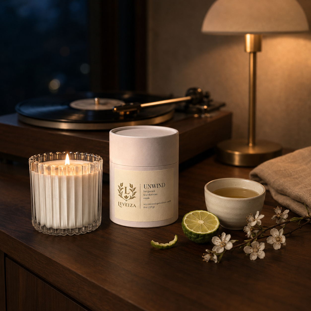 Lit Unwind candle and tube with evening tea, bergamot and tea blossoms beside a record player in warm lamplight.
